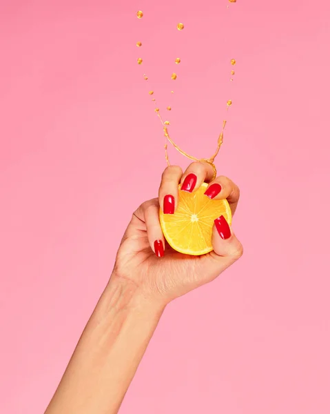 Vitamines. Female hand with bright manicure squeezes half of orange over pink background. Pop art food photography. Drops of juice fly up. Concept of healthy eating, art, fashion, creativity and ad