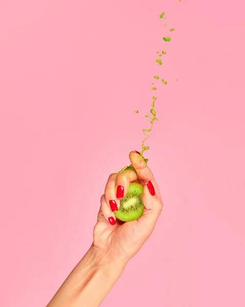 Authentic female hand squeezes half of kiwi fruit over pink background. Pop art food photography. Drops of juice fly up. Breaking laws of physics. Concept of healthy eating, art, fashion, ad
