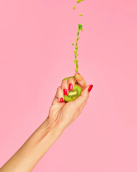 Ripe and juicy. Authentic female hand squeezes half of kiwi fruit over pink background. Pop art food. Drops of juice fly up. Breaking laws of physics. Concept of healthy eating, art, fashion, ad