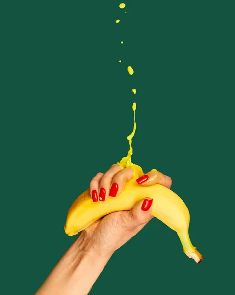 Tropical taste. Hand with bright manicure squeezes banana over green background. Pop art food photography. Drops of juice fly up. Concept of healthy eating, art, fashion, creativity and ad
