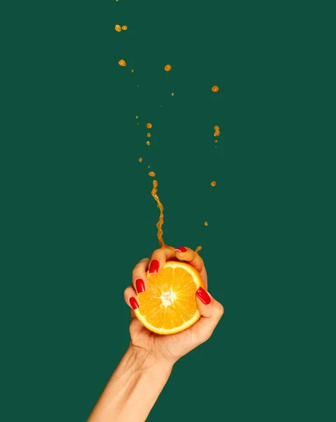 Vitamines. Female hand with bright manicure squeezes half of orange on green background. Pop art food photography. Drops of juice fly up. Concept of healthy eating, art, fashion, creativity and ad