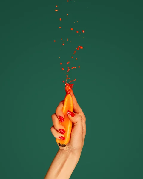 Elegant womans hand squeezes carrot with force and drops of juice fly up on green background. Pop art photography. Concept of fashion, creativity, surrealism. Healthy eating, ecology products