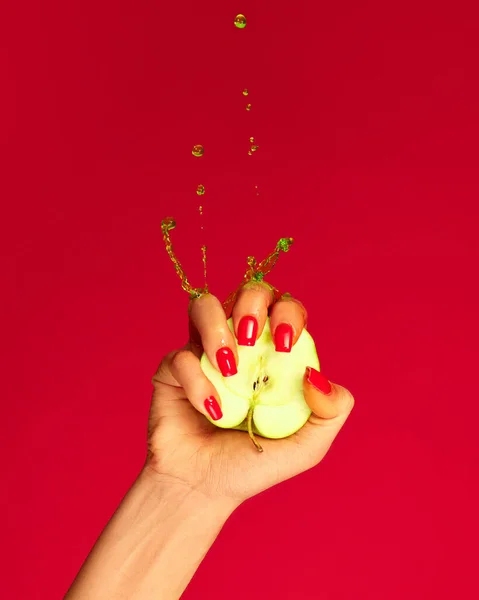 Closeup female hand with red painted nails squeezing half of apple fruit over red background. Contemporary art. Drops of juice fly up. Breaking laws of physics. Concept of healthy eating, fashion, ad