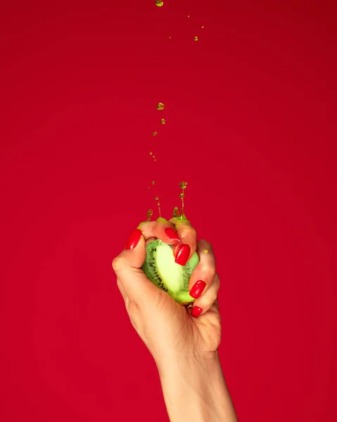 Ripe and juicy. Authentic female hand squeezes half of kiwi fruit over red background. Pop art food. Drops of juice fly up. Breaking laws of physics. Concept of healthy eating, art, fashion, ad