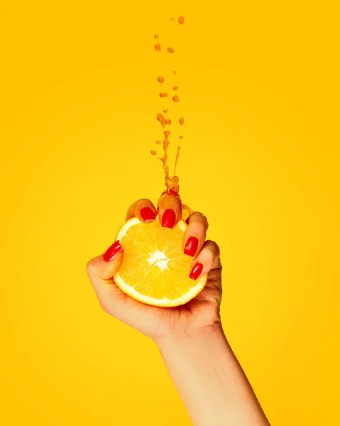 Summer vitamines. Hand with red manicure squeezes half of orange over yellow background. Pop art food photography. Drops of juice fly up. Concept of healthy eating, art, fashion, creativity and ad
