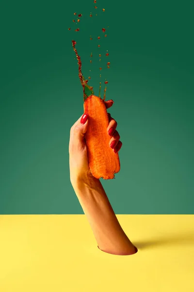 Elegant womans hand squeezes carrot with force and drops of juice fly up on green yellow background. Pop art photography. Concept of fashion, creativity, surrealism. Healthy eating, ecology products