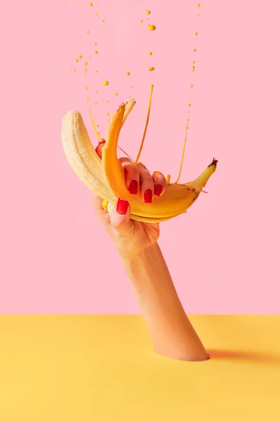 Female hand with bright manicure squeezes banana over pink-yellow background. Pop art food photography. Drops of juice fly up. Concept of healthy eating, art, fashion, creativity and ad