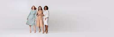 Full-length portrait of diverse beautiful young women in elegant dresses standing together over light background. Concept of spring mood, lightness, beauty, fashion, carefree. Banner for ad