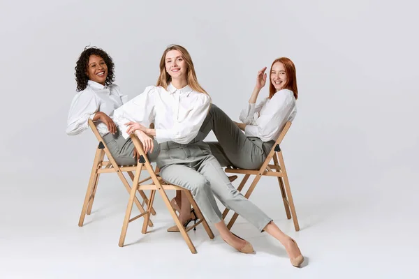 Businesswomen. Portrait of adorable stylish young women in office style clothes sitting on chairs over light background. Concept of beauty, fashion, business, ad. Models look happy