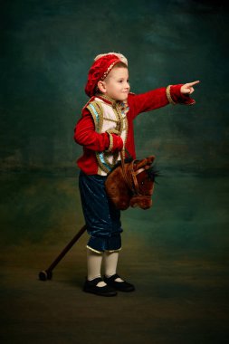 One cute little boy dressed up as medieval little prince and pageboy ride toy horse over dark vintage style background. Vintage fashion, emotions, theater art concept. Eras comparison