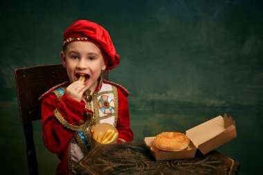 Happy cheerful little boy wearing costume of medieval page boy and prince eating hamburger with french fries over dark green background. Fast food, happiness, childhood and tastes concept