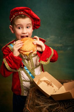 Happy cheerful little boy wearing costume of medieval page boy and prince eating hamburger with french fries over dark green background. Fast food, happiness, childhood and tastes concept