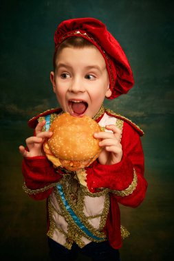 Happy cheerful little boy wearing costume of medieval page boy and prince eating hamburger over dark green background. Fast food, happiness, childhood and tastes concept