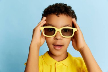 Surprised little african boy in stylish sunglasses and yellow summer shirt looking at camera over blue background. Concept of facial expression, happiness, kids emotions. Shock, wow