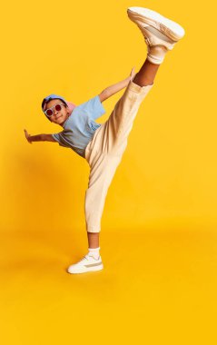 Küçük Afrikalı bir çocuğun dinamik portresi, şık sokak kıyafetleri içinde hip-hop dansçısı parlak sarı arka planda dans ediyor. Müzik kavramı, dans mutluluğu. Çocuk mutlu ve sportif görünüyor.