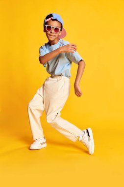 Dynamic portrait of little african boy, hip-hop dancer in stylish street style clothes dancing over bright yellow background. Concept of music, dance happiness. Kid looks happy and sportive