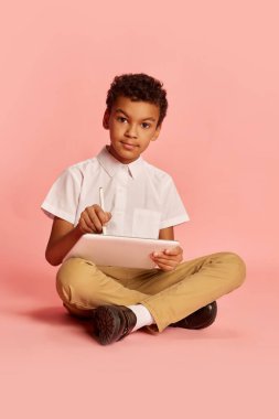 Portrait of school age american-african boy, pupil in modern school uniform sitting with tablet and drawing over pink background. Concept of new generation, education, studying and fashion.