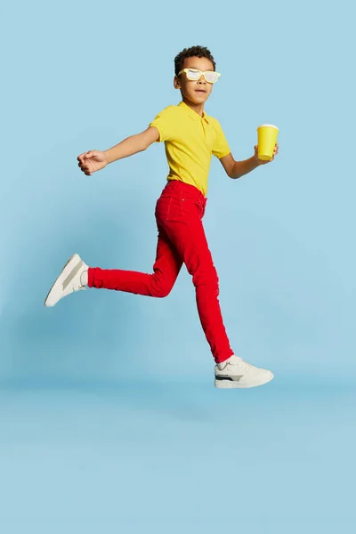 Run away. Fashionable little african boy in stylish sunglasses and yellow summer shirt and red trousers posing over blue background. Concept of fashion, happiness, kids emotions