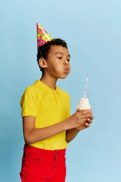 Make wish. School age boy in bright colors clothes with birthday cone and cake isolated on blue background. Concept of kids fashion, emotions, art photography, style, holidays, dreams, ad