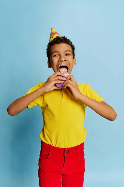 Delighted school age boy in bright colors clothes with birthday cone eating sweet cake isolated on blue background. Concept of fashion, art photography, style, holidays, dreams, ad. Happiness