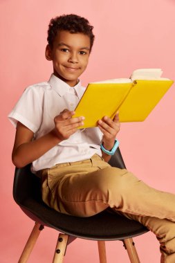 Closeup of school age smiling boy, pupil in modern school uniform sitting with book over pink background. Concept of new generation, education, studying and fashion.