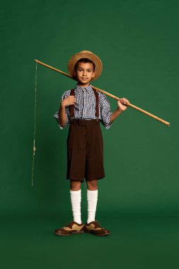 Fisherman. Cute handsome little boy, child in checkered shirt and suspender shorts isolated over dark green background. Concept of childhood, friendship, retro fashion style