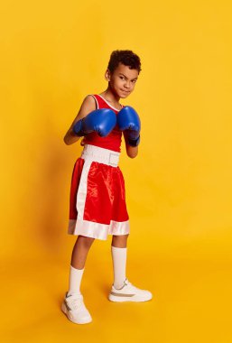 Champion. Active sportive little boy, beginner boxer in sports uniform and boxing gloves isolated over yellow background. Concept of sport, active and healthy lifestyle, kids emotions
