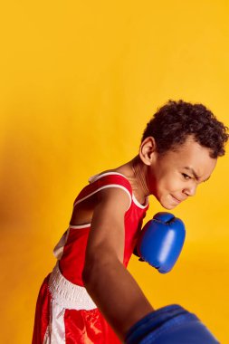 Punching. Active sportive little boy, beginner boxer in sports uniform and boxing gloves isolated over yellow background. Concept of sport, active and healthy lifestyle, kids emotions