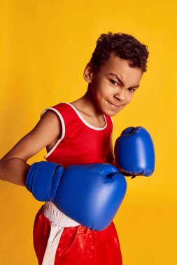 Challenge. Closeup of sportive little boy, beginner boxer in sports uniform and boxing gloves isolated over yellow background. Concept of sport, active and healthy lifestyle, kids emotions