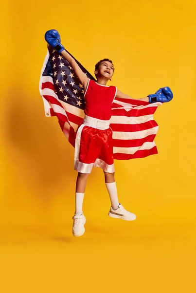 Winner emotions. Little male beginner boxer in sports uniform and boxing gloves standing with national american flag isolated over yellow background. Concept of sport, kids emotions, success, win