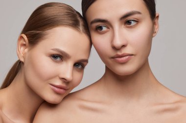 Tenderness. Closeup portrait of two adorable young girls with well-kept skin posing over grey background. Couple of models with bare shoulders. Concept of beauty, spa, skin care and health