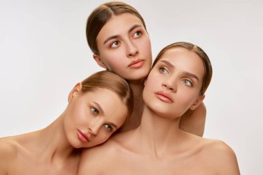 Beauty. Three beautiful young girls with well-kept skin posing over grey background. Fashion models with bare shoulders. Concept of care, spa, skin care and healthy lifestyle