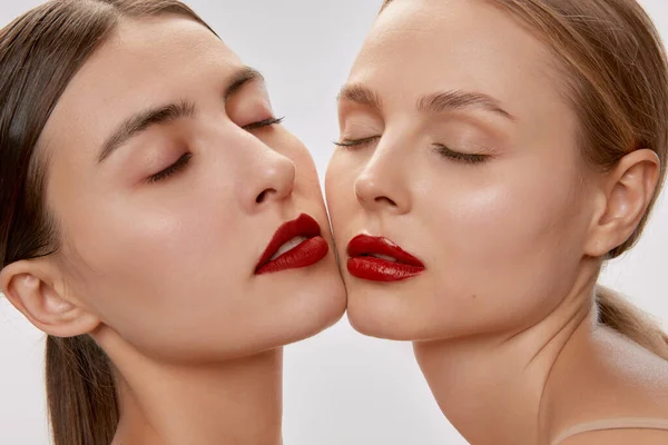 Light touch. Two sensual beautiful young women with nude makeup and red lipstick posing with closed eyes. Concept of love, emotions, beauty, vision, health and fashion