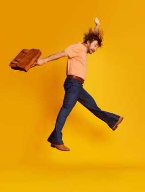 Excited mature bearded hairy man, hippie in stylish sunglasses and flared jeans running jumping over yellow background. Retro fashion style, american culture, emotions and ad