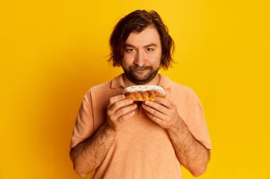 Half-length portrait of bearded man looking at camera and tasting sugar bun isolated over yellow background. Concept of facial expression and human emotions.