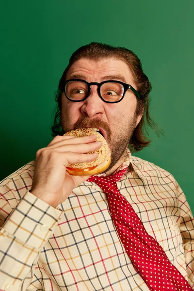 Fast food taste. Retro mature man with moustache and beard in eyeglasses in vintage fashion clothes tasting hamburger over dark green background. Concept of retro style, emotions, business, work