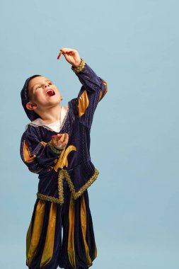 Jöleli şeker yiyorum. Orta çağ çocuğu kostümlü küçük mutlu çocuk, açık mavi arka planda küçük prens. Çocuk duyguları kavramı, dönem karşılaştırması, moda, tiyatro, sanat, festival