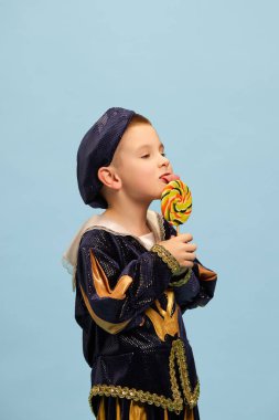 Şekerler. Ortaçağ şamatacı kostümlü sevimli küçük çocuk, açık mavi arka planda büyük lolipoplu küçük prens. Çocuk duyguları kavramı, dönem karşılaştırması, moda, tiyatro