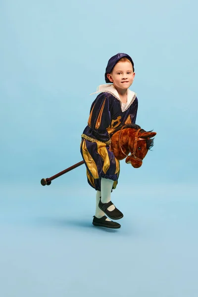 快乐可爱的小男孩打扮成中世纪的小王子和拉皮条的男孩骑在浅蓝色背景的玩具马背上 老式时尚 戏剧艺术理念 Eras比较 — 图库照片
