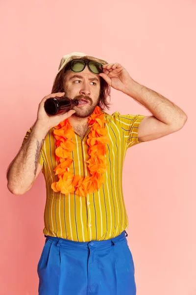 Surprise. Bearded man in summer shirt and short with Hawaiian flower garland on chest tasting beer over pink background. Leisure activity, vacation, summer, rest and drinks concept