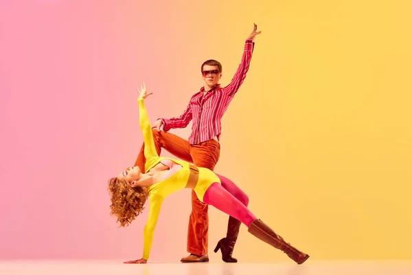 Disco. Young stylish emotional man and woman, professional dancers in retro style clothes dancing disco dance over pink-yellow background. 1970s, 1980s fashion, music concept