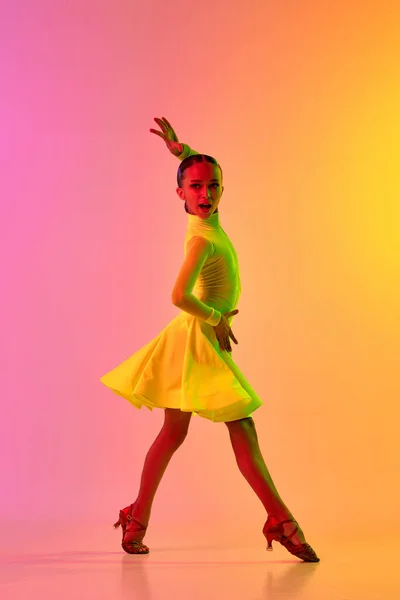 Elegant little girl in adorable stage outfit, dress dancing ballroom dance over gradient pink-yellow background in neon light filter. Concept of beauty, professional dances, skills. Emotions in dance