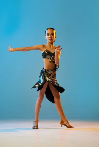 Rumba dance. Beautiful little girl in stage attire dancing latino madern ballroom dance over blue background. Music, dance, education, professional dance school concept. Copy space for ad