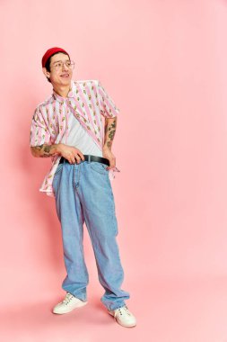 Tarz sahibi bir adamın uzun bir fotoğrafı, kemerini bağlayan ve pembe arka plana bakan gülümseyen genç bir adam. Moda anlayışı, gençlik, öğrenim hayatı