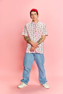 Sakinlik. Dövmeli, elleri çapraz duran ve pembe arka planda kameraya bakan şık bir adamın tam boy fotoğrafı. İnsan duyguları, moda, gençlik, öğrenci hayatı kavramı