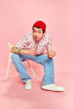 Şaşırdım. Modaya uygun bir adam pembe arka planda otururken öne eğilip poz veriyor. Duygular, moda, gençlik, öğrenci hayatı. Kot pantolon, şapka ve gömlek giyen manken.