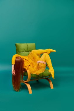 Koltuğunda poz veren kızıl saçlı kızın yaratıcı portresi. Cyan renkli arka planda garip pozlar veriyor. Kişisel olmayan duygular, vücut dili. Moda, güzellik, akıl sağlığı.