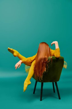 Creative portrait of redhead girl with long straight hair posing on armchair, sitting in strange poses over cyan color background. Impersonal emotions, body language. Fashion, beauty, mental health clipart