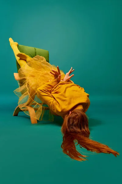 stock image Creative portrait of redhead girl with long straight hair posing on armchair, sitting in strange poses over cyan color background. Impersonal emotions, body language. Fashion, beauty, mental health