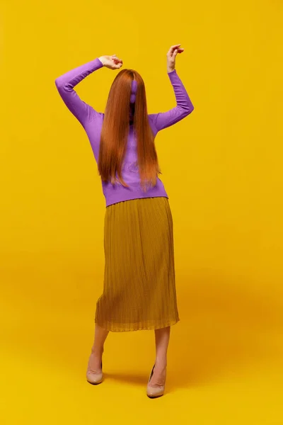 Weird dance. Faceless portrait of slim woman with long straight red hair wearing retro fashion outfit in action over yellow studio background. Impersonal emotions, body language